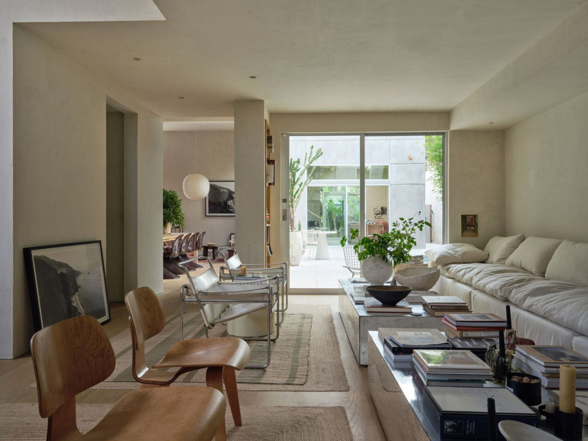 On the opposite side of the front door, the living room features a long, overstuffed sofa that stretches across the plaster wall, while layered jute rugs and two large coffee tables are stacked with art books.