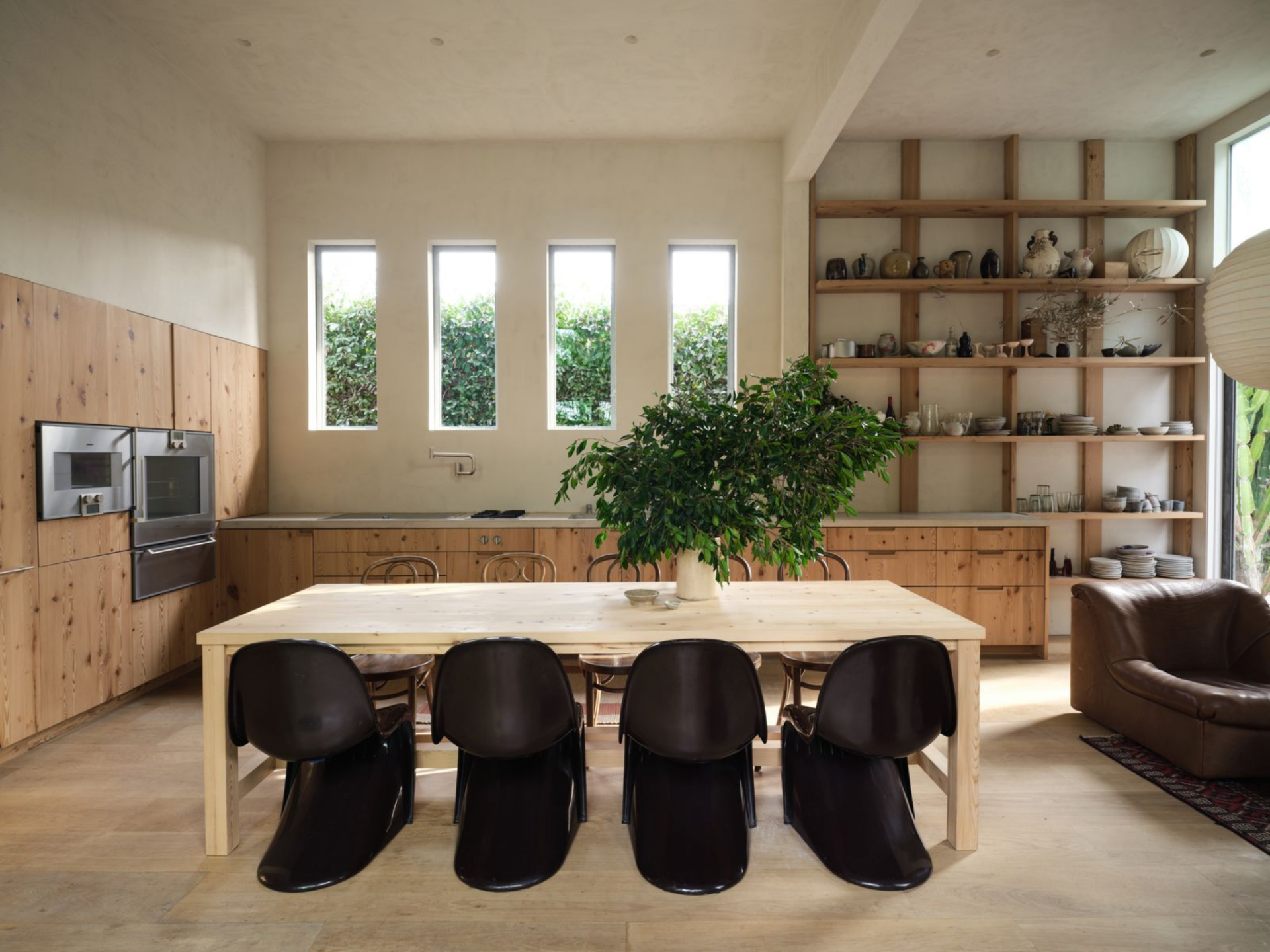 Lastly, the center of the room holds a large, natural wood dining table surrounded by sculptural dark chairs on one side and wood ones on the other, combining modern and rustic elements.