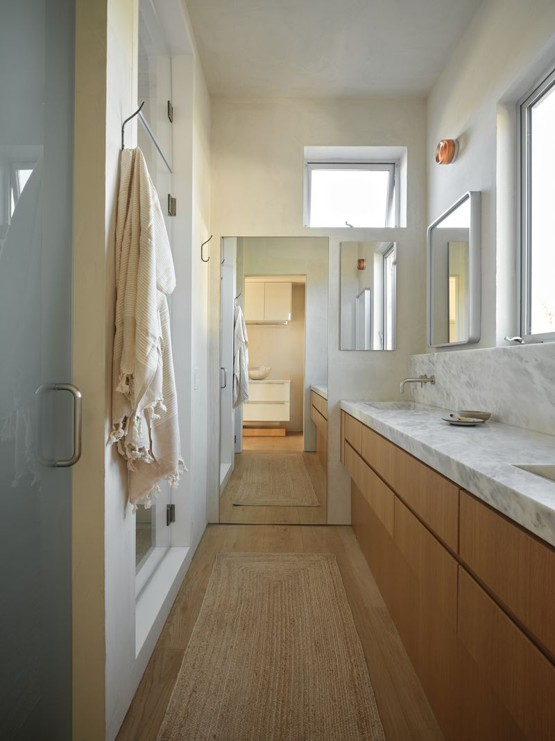 The conected bath stretches vertically with a long marble countertop lining the wall on top of warm wood cabinetry. 