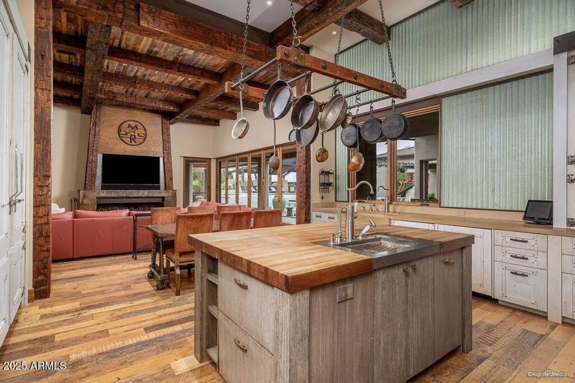 At the center sits a large butcher-block island with an integrated sink and open shelving, topped by a suspended pot rack that keeps cast-iron pans and copper cookware within easy reach.