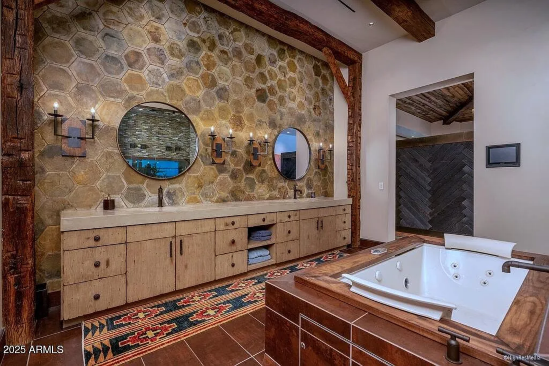 From there, the suite flows into a spa-like bathroom wrapped in rich materials. A long double vanity stretches beneath a striking honeycomb-patterned stone wall, with round mirrors and iron sconces.