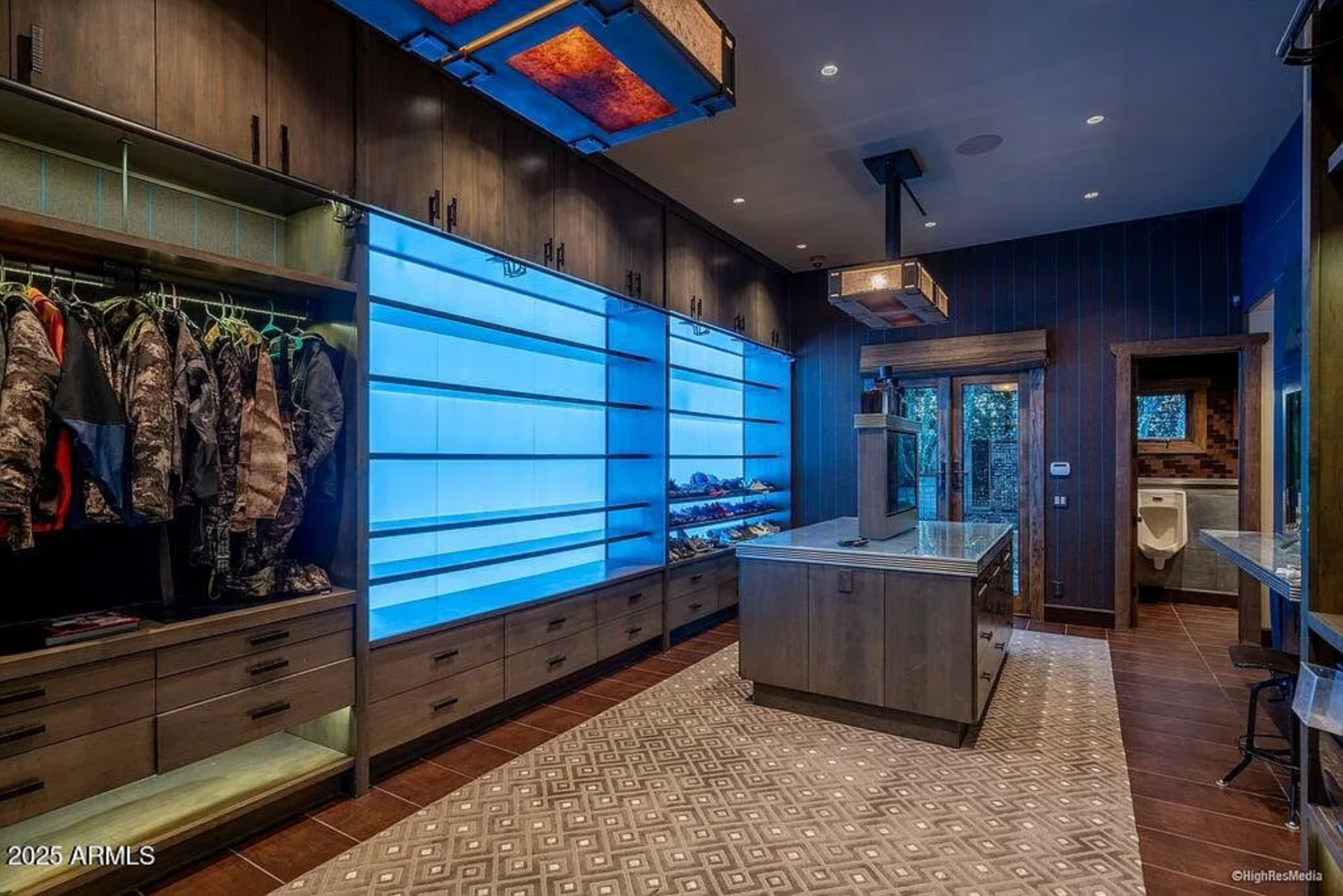 The "his" closet is finished in darker tones with custom cabinetry, illuminated display shelving, and a central island for storage and folding.