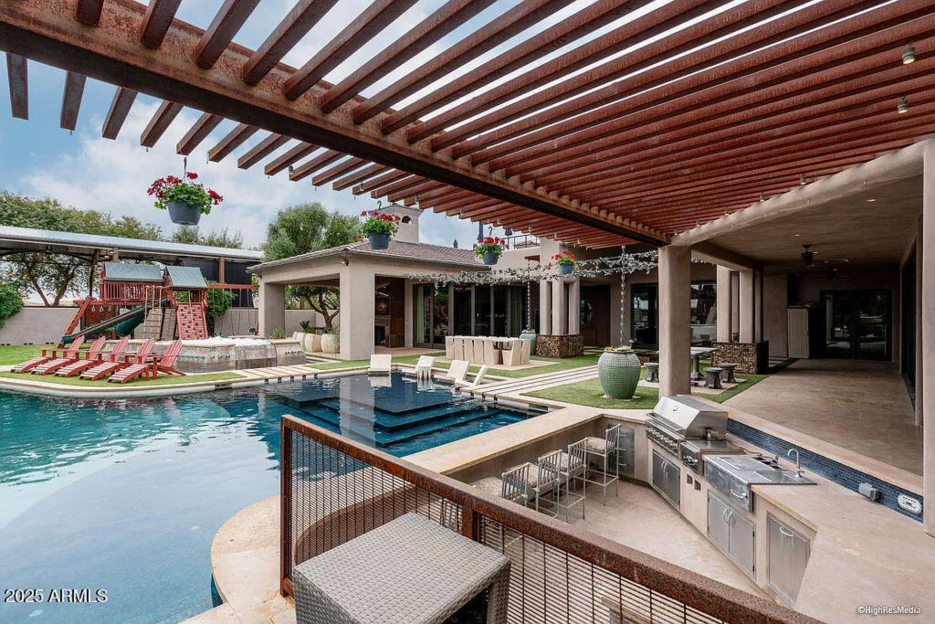 Just beyond the main house, a covered patio with a slatted wood pergola stretches along the back, creating a shaded lounge complete with a full outdoor kitchen, built-in grill, bar seating, and dining areas that flow directly from the interior living spaces.
