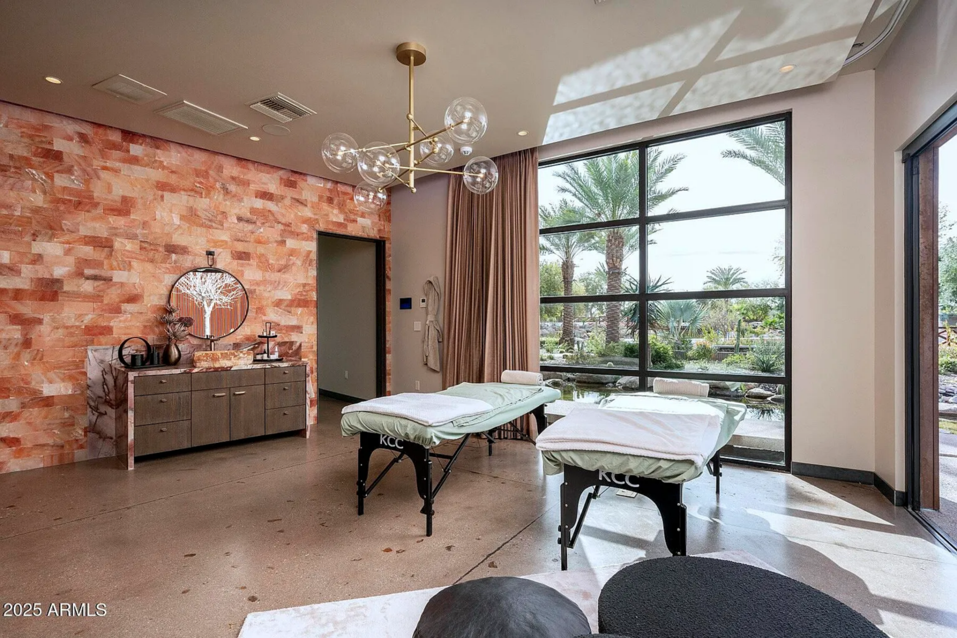 The massage room features two professional treatment tables positioned beneath a sculptural chandelier, with a full wall of windows overlooking the landscaping.