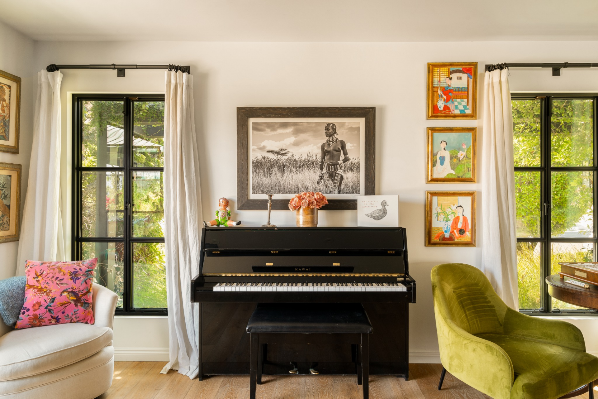 A black upright piano, sitting between tall windows dressed in soft white curtains, framed by plush chairs. 