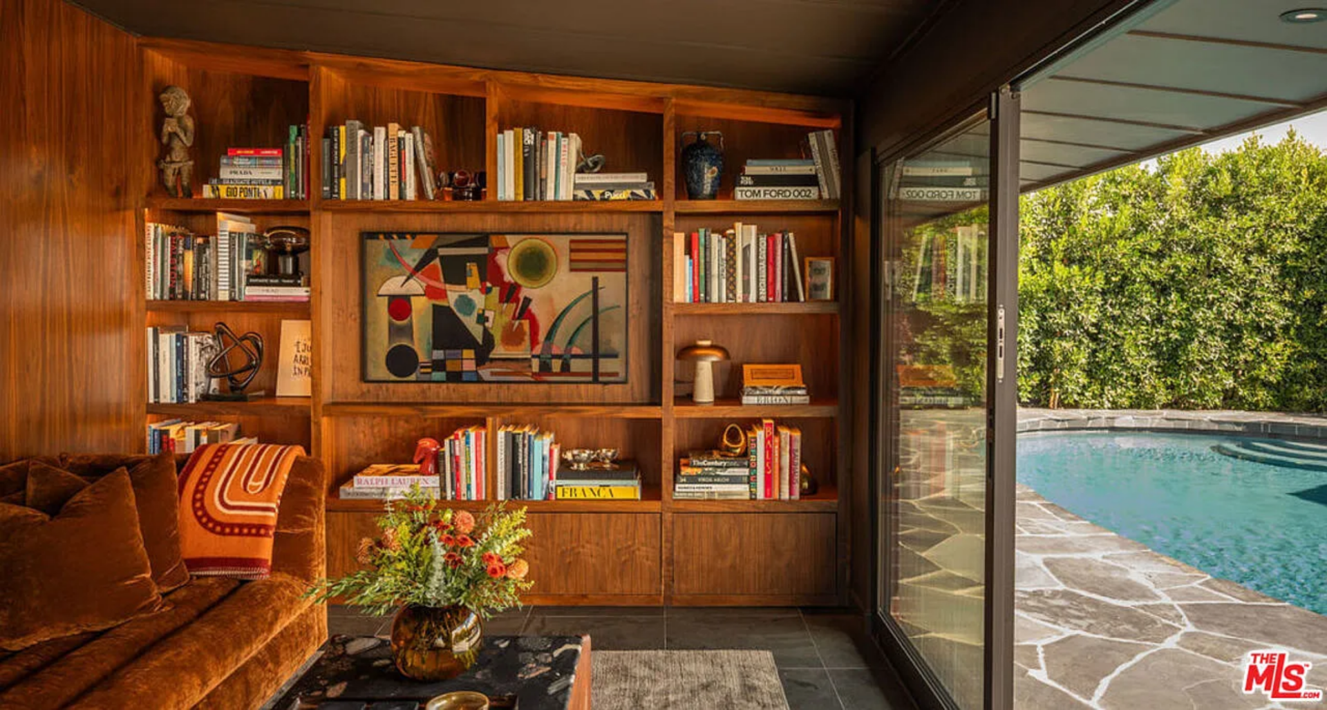Tucked behind the slatted partition, the nook is wrapped in warm wood paneling and built-in shelving, softly lit to showcase a variety of books, sculptures, and framed artwork next to a deep, curved velvet sofa.