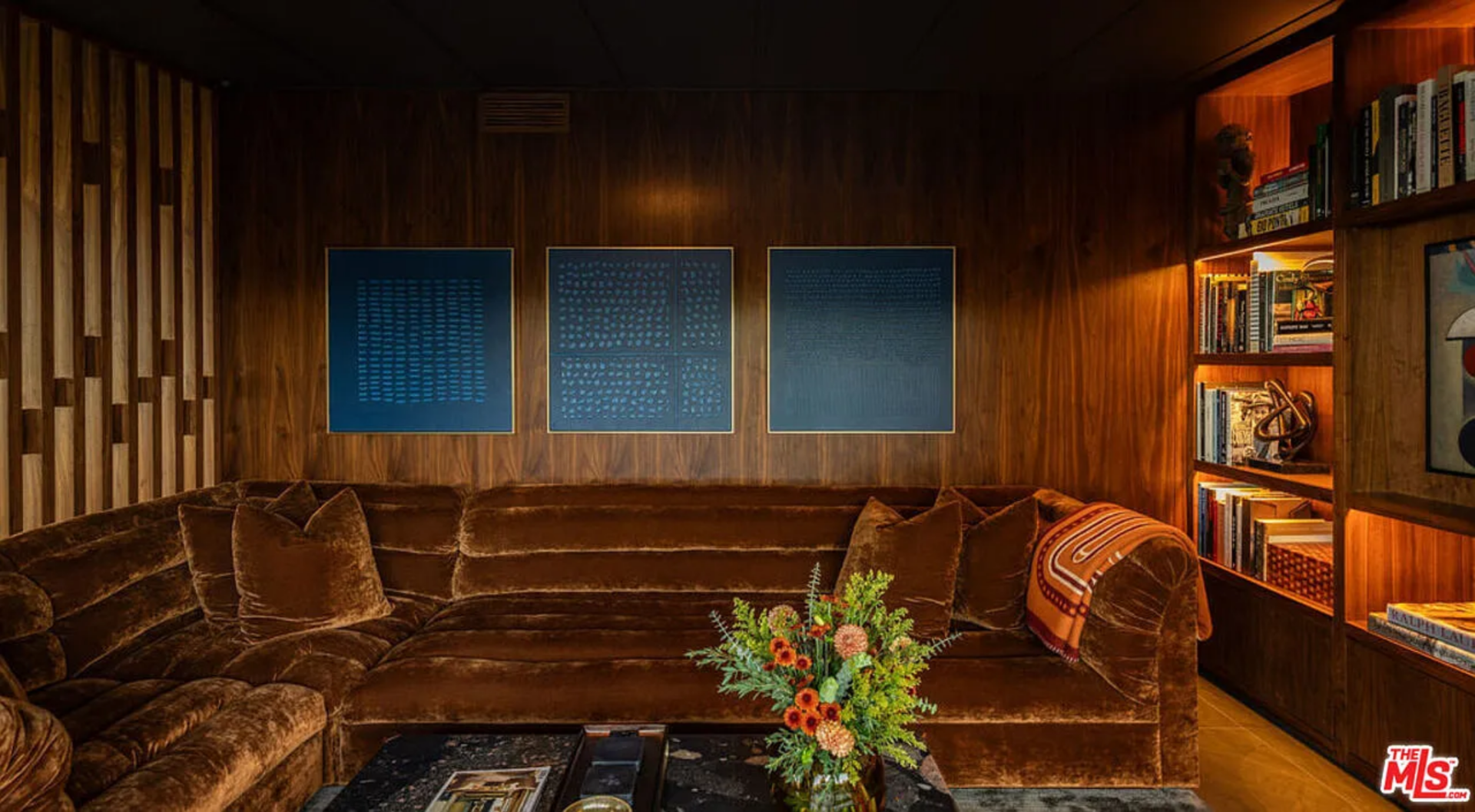 Tucked behind the slatted partition, the nook is wrapped in warm wood paneling and built-in shelving, softly lit to showcase a variety of books, sculptures, and framed artwork next to a deep, curved velvet sofa.