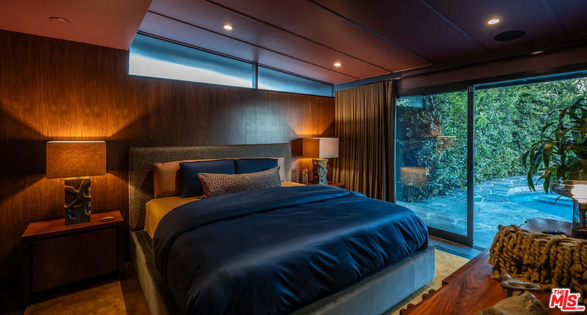 In a corner of the house, the primary suite features a low-profile upholstered bed centered against the paneled wall, framed by sculptural table lamps that cast warm, focused light across the grain along the wall.