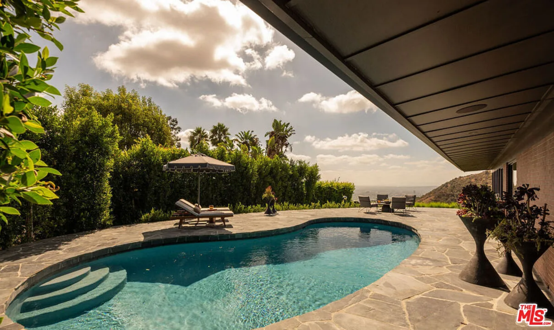 A freeform pool sits in the center of the terrace, its curved edge and built-in entry steps softening the linear roofline overhead, while lounge chairs and a shaded umbrella are positioned along the perimeter.