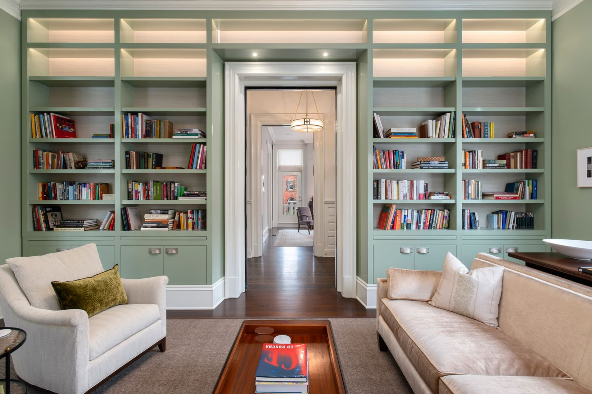 Directly to the left of the entry hall, past the front door, the library is lined with floor-to-ceiling built-ins painted a muted sage, wrapping the doorway in open shelving above and closed cabinetry below.