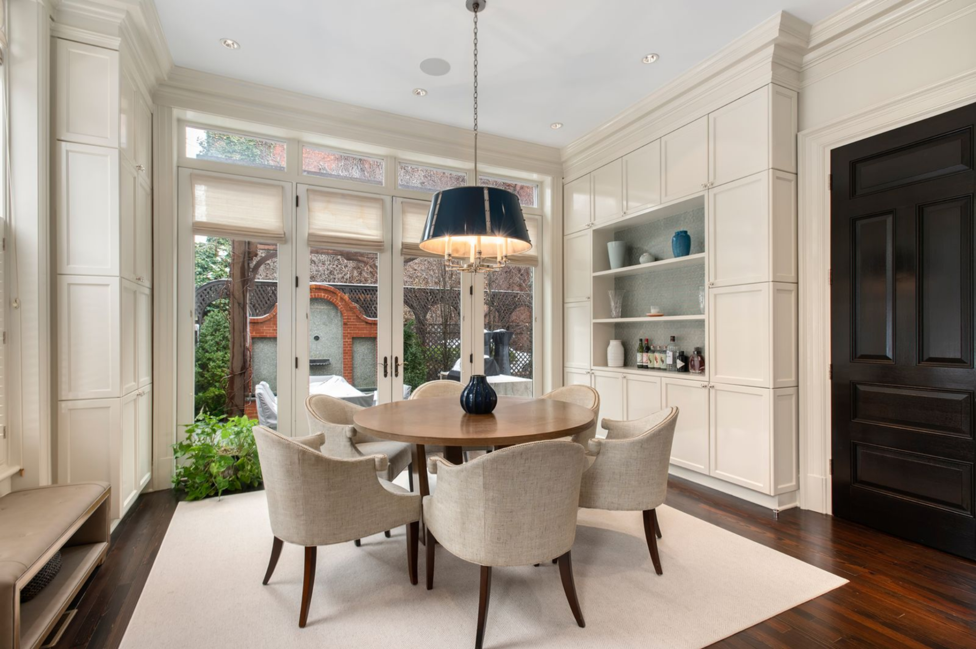 A round wood table is centered beneath a tailored pendant fixture, surrounded by upholstered chairs and bordered by built-in storage and display shelving. 
