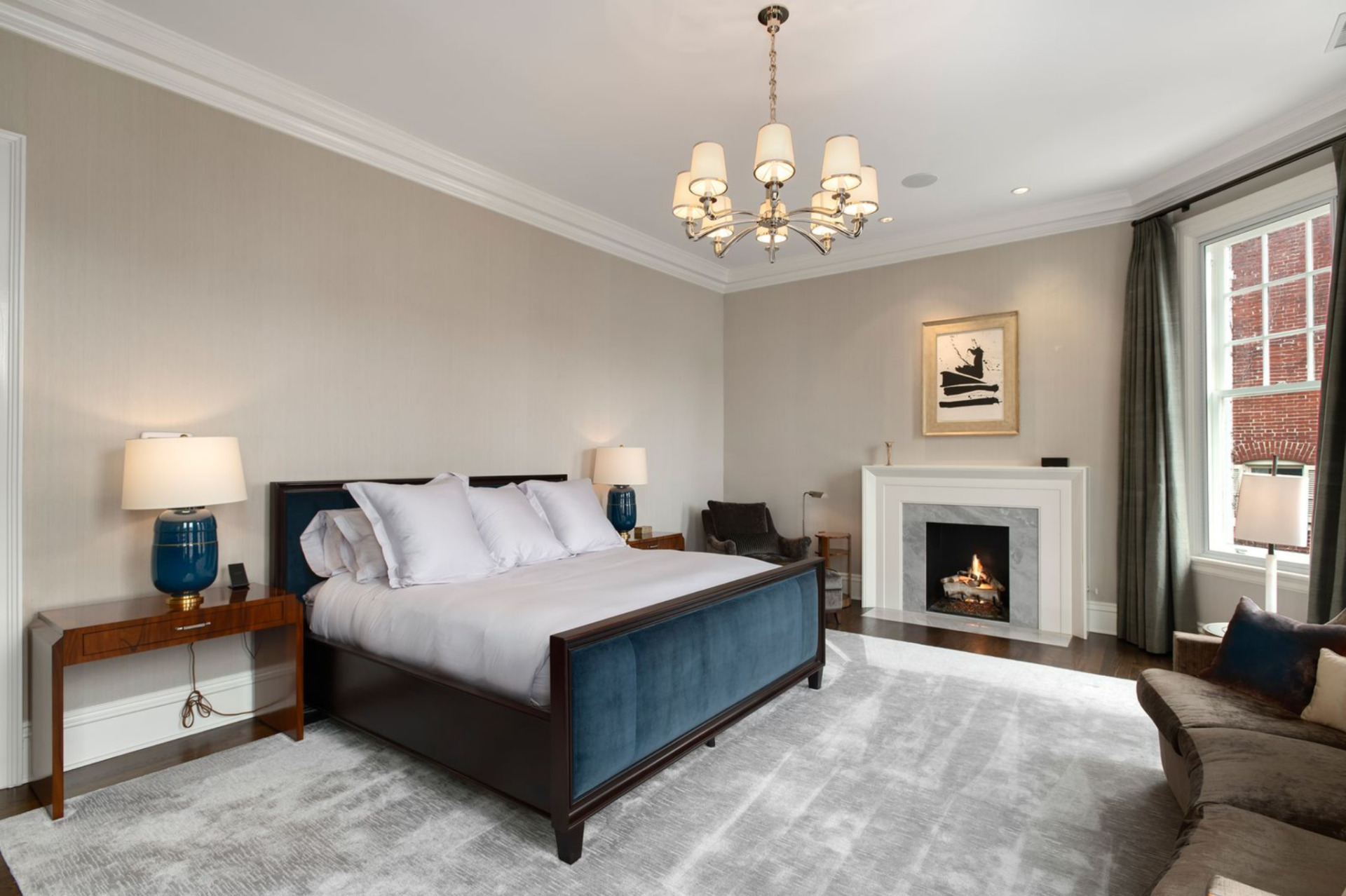 Inside the primary suite, the bedroom is centered around a tailored bed framed in dark wood with upholstered panels, while a gas fireplace with a marble surround sits beneath a framed work of art. 