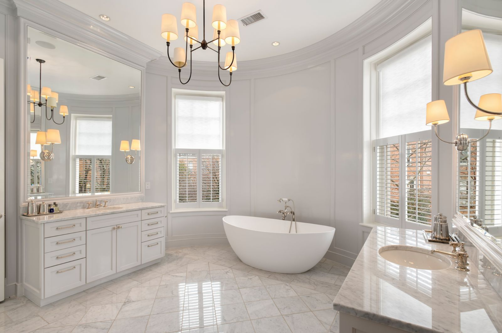 Dual vanities with polished stone countertops line opposite walls beneath oversized mirrors, while a freestanding soaking tub is positioned between tall windows fitted with shutters. 
