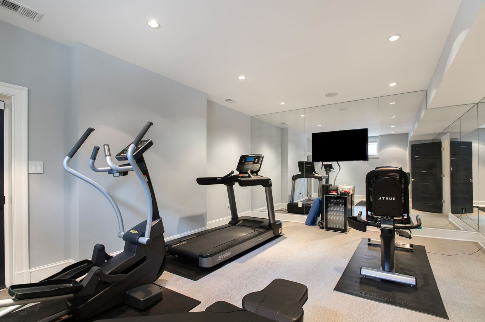 In a separate area, the home gym is filled with professional-grade equipment, including a treadmill, elliptical, stationary bike, and bench, each set on protective mats.