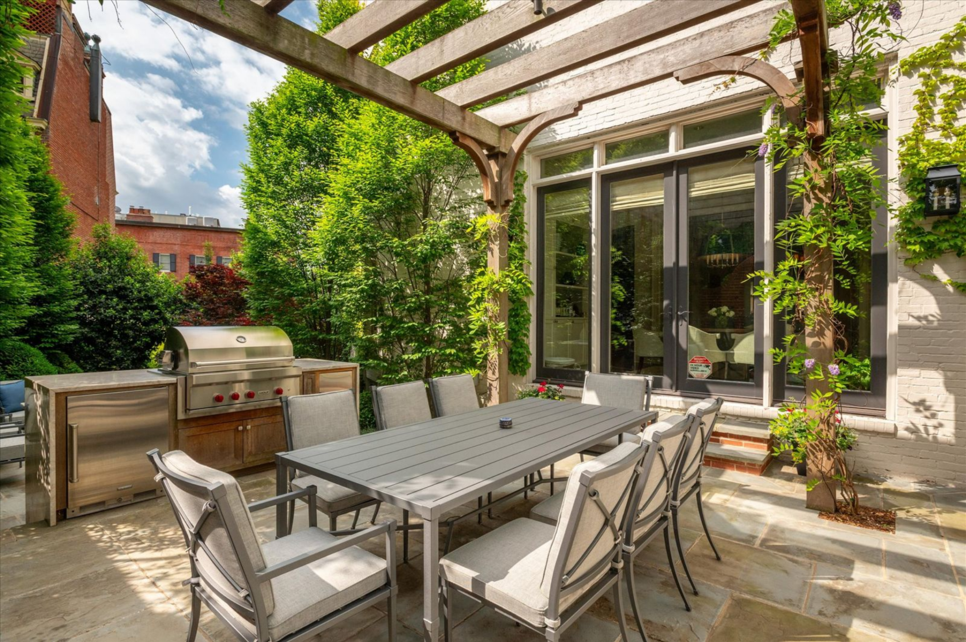 A stone-paved dining terrace sits beneath a wood pergola, where a long rectangular table is positioned next to a built-in grill station.
