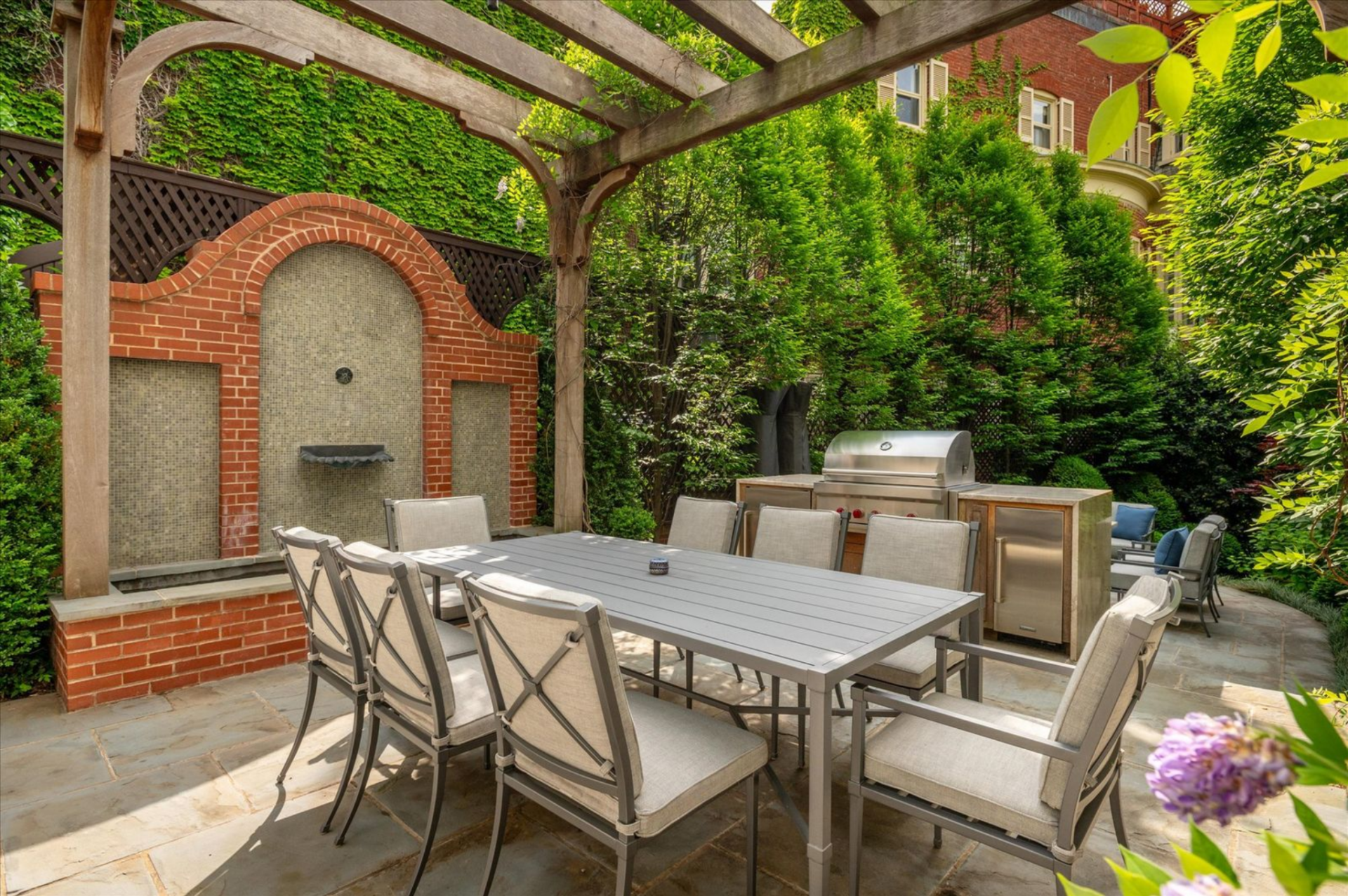 Opposite the dining area, a brick fountain with a mosaic-tiled inset serves as a focal point, framed by manicured hedges and layered plants that enclose the courtyard in privacy.