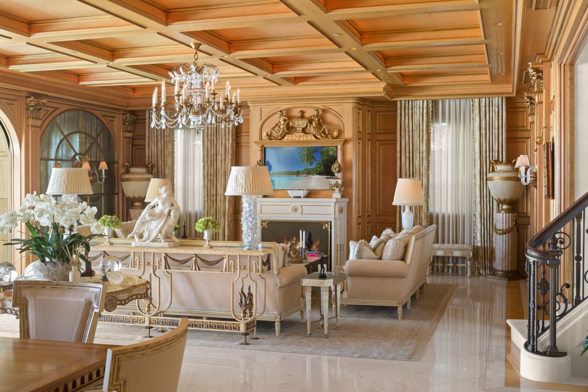The guest house living room opens beneath a coffered ceiling designed with hand-painted panels, each inset framed by rich wood detailing, and has crystal chandeliers hanging down.