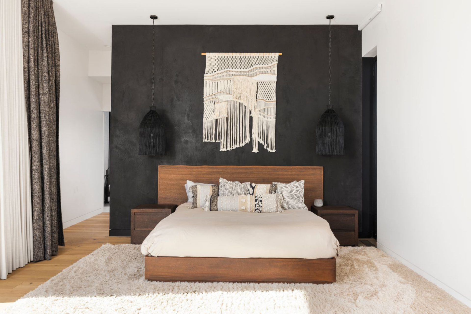 The primary suite occupies its own wing of the home, beginning with a calm, minimalist bedroom featuring a warm wood bed frame and a dramatic dark accent wall. 