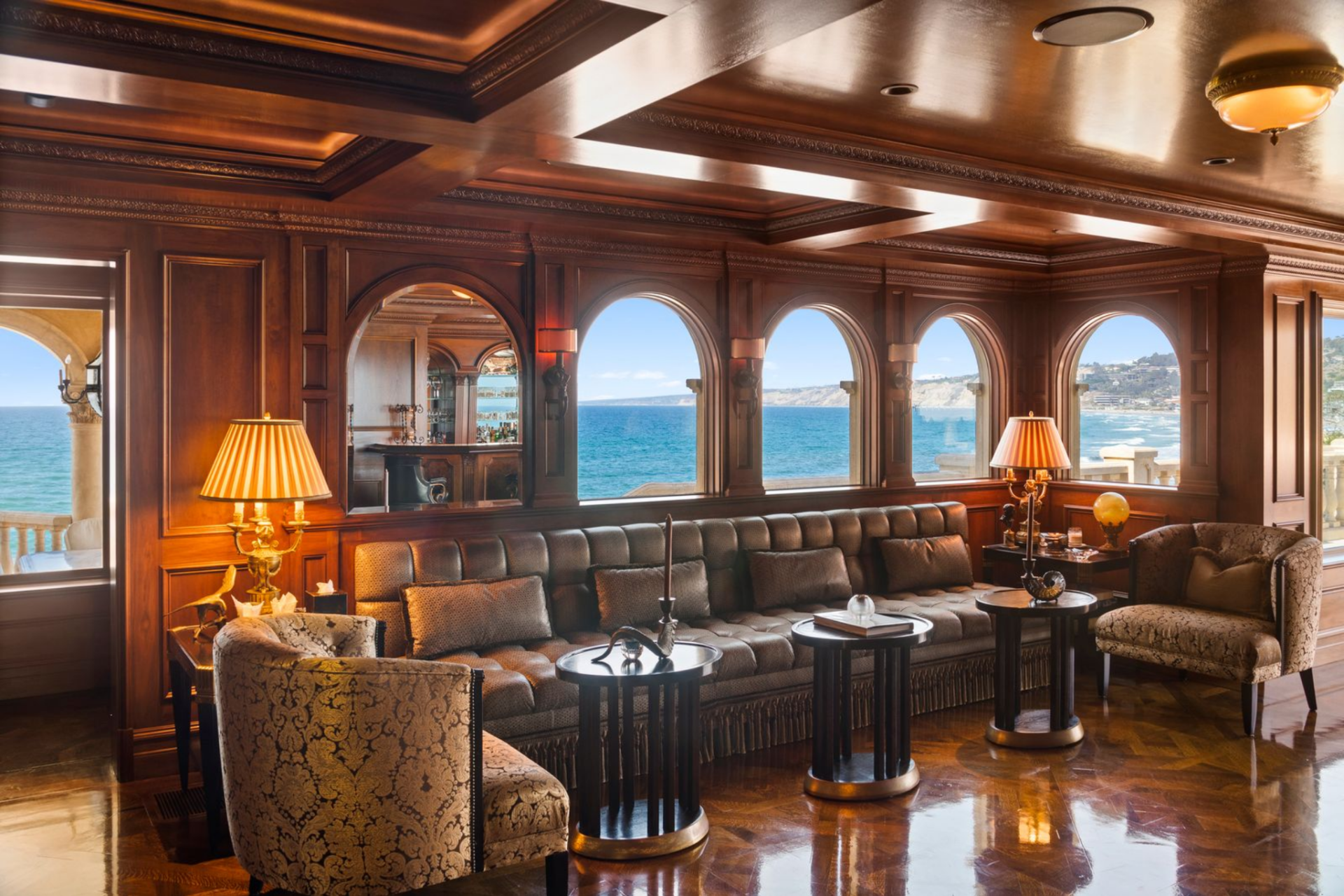 Beyond the bar, a lounge area features tufted leather seating, patterned armchairs, and dark round tables positioned beneath a series of arched windows framing uninterrupted ocean views. 