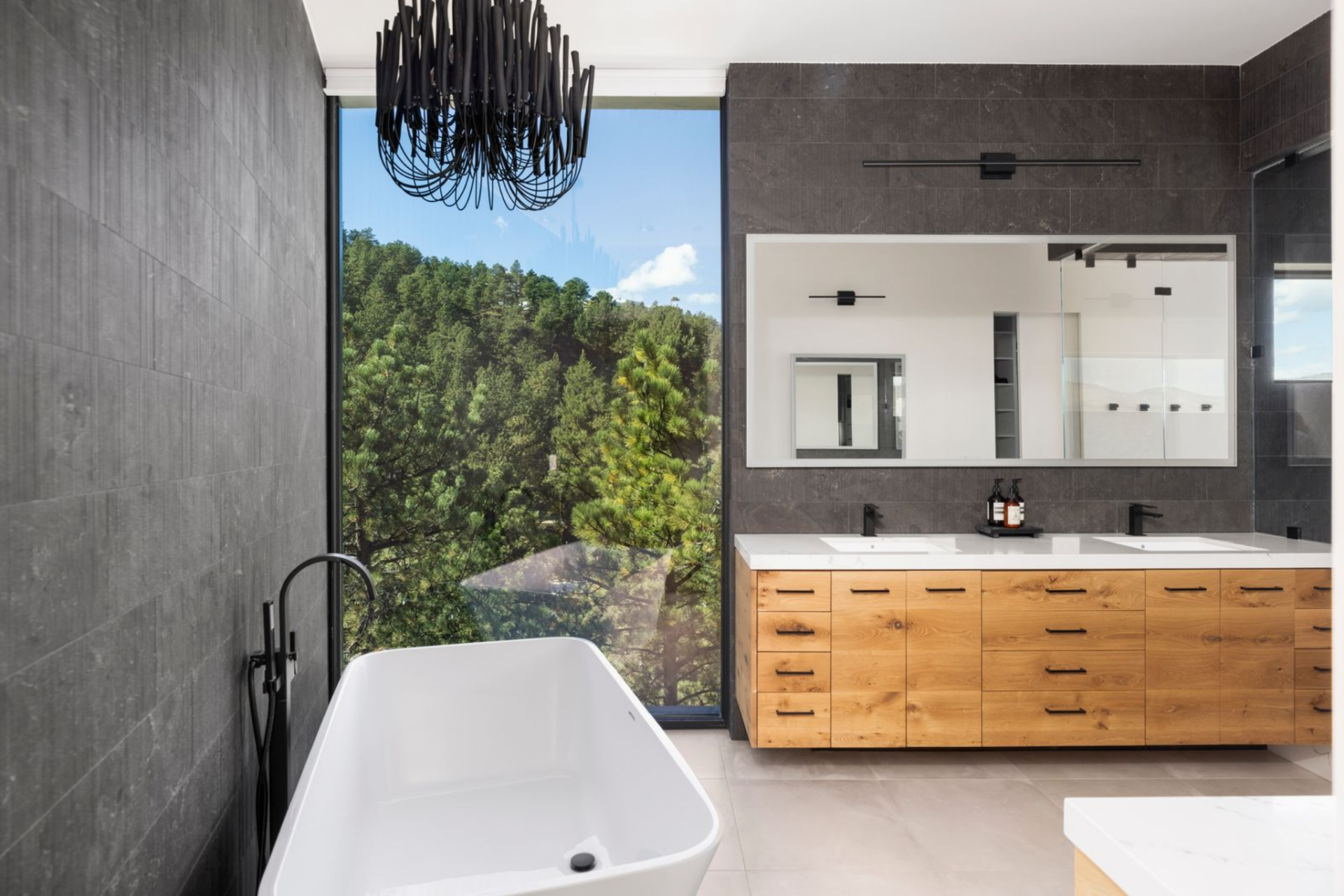 The en-suite bathroom has a double vanity with wood cabinetry sitting opposite a glass-enclosed shower and soaking tub, both framed by textured stone and oversized windows that pull in forest views.