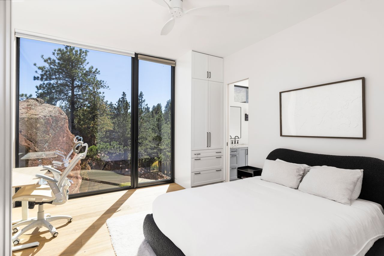 This bedroom is bright with clean white walls, light wood floors, and a large glass wall that pulls in natural light and frames the surrounding rock and trees just outside. 