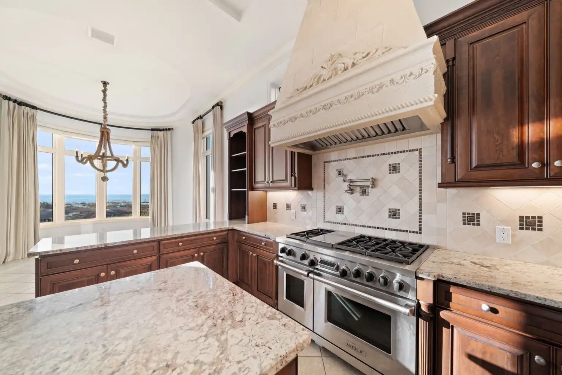 A professional-grade gas range is framed by a detailed stone hood and custom tile backsplash, while integrated appliances and deep storage keep the room clean.