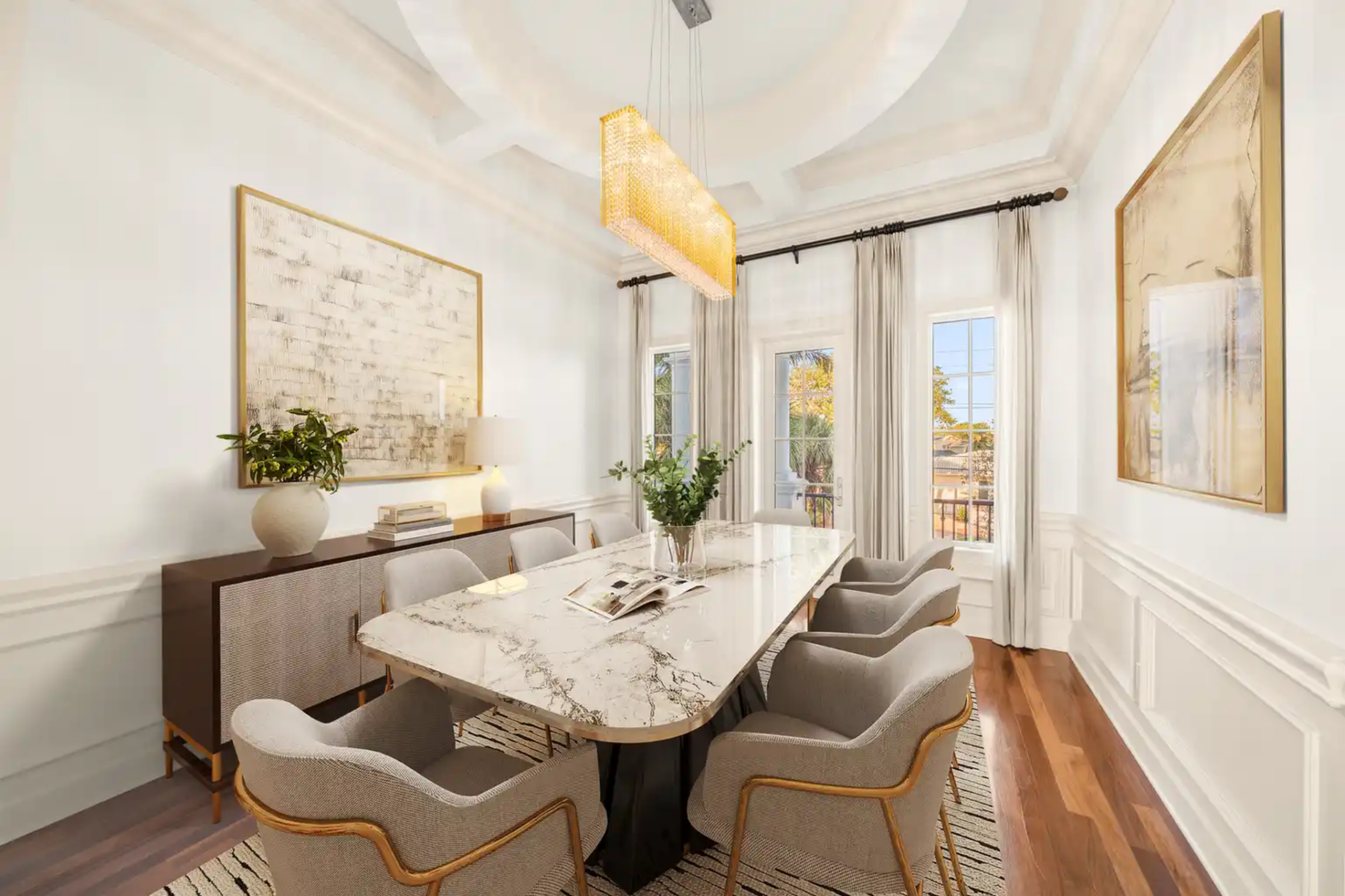 The formal dining room features a long marble-topped table that sits beneath a linear chandelier, its warm glow reflecting off crisp white walls and paneled millwork.