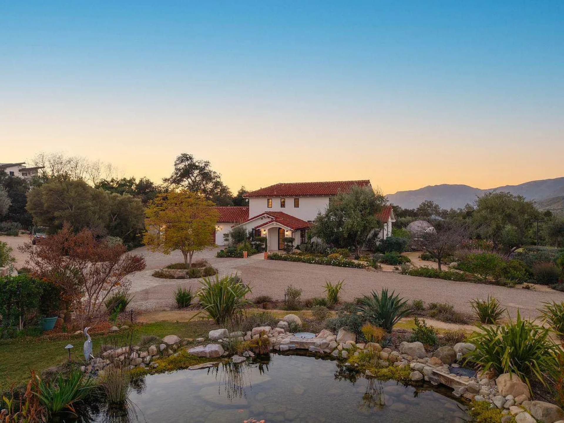 The main house sits beyond a gravel drive, framed by mature trees, native plants, and a still pond that reflects soft light.