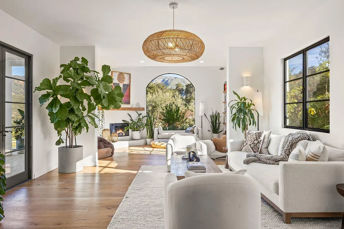 Soft plaster walls, black steel–framed windows, and layered natural textures have sunlight washing over them drawing your eye toward the arched opening and fireplace.