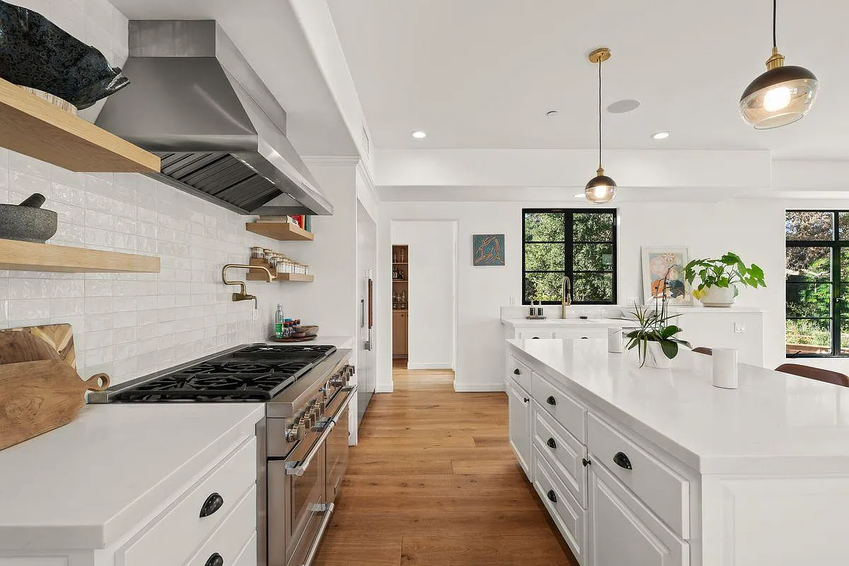 White cabinetry runs wall to wall, broken up by open wood shelves, a substantial stainless-steel range and hood, and more black-framed windows that pull in views of the surrounding greenery.