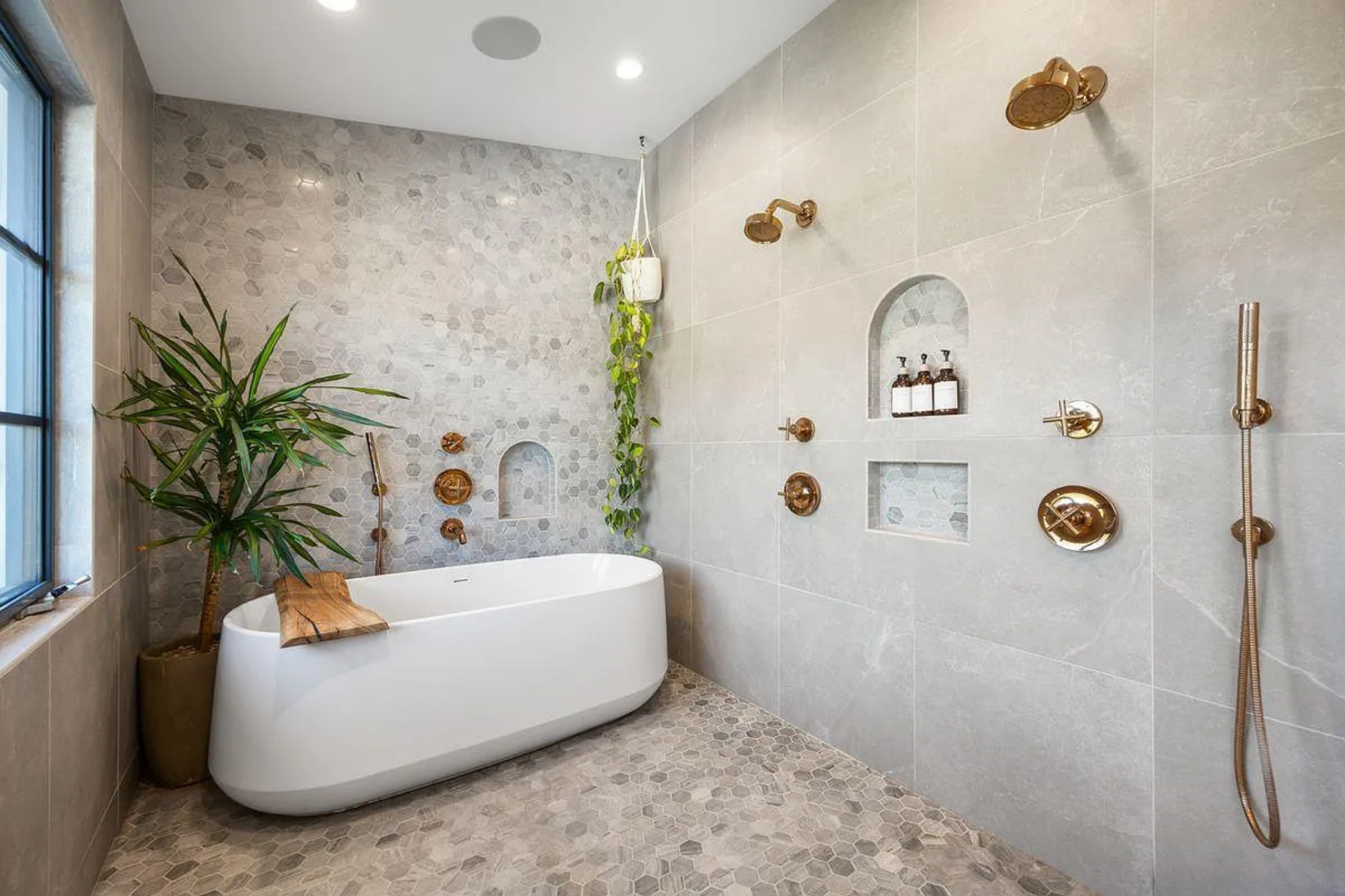 Across the room, a freestanding soaking tub is set against a wall of stone and patterned tile, framed by arched niches holding bath essentials.