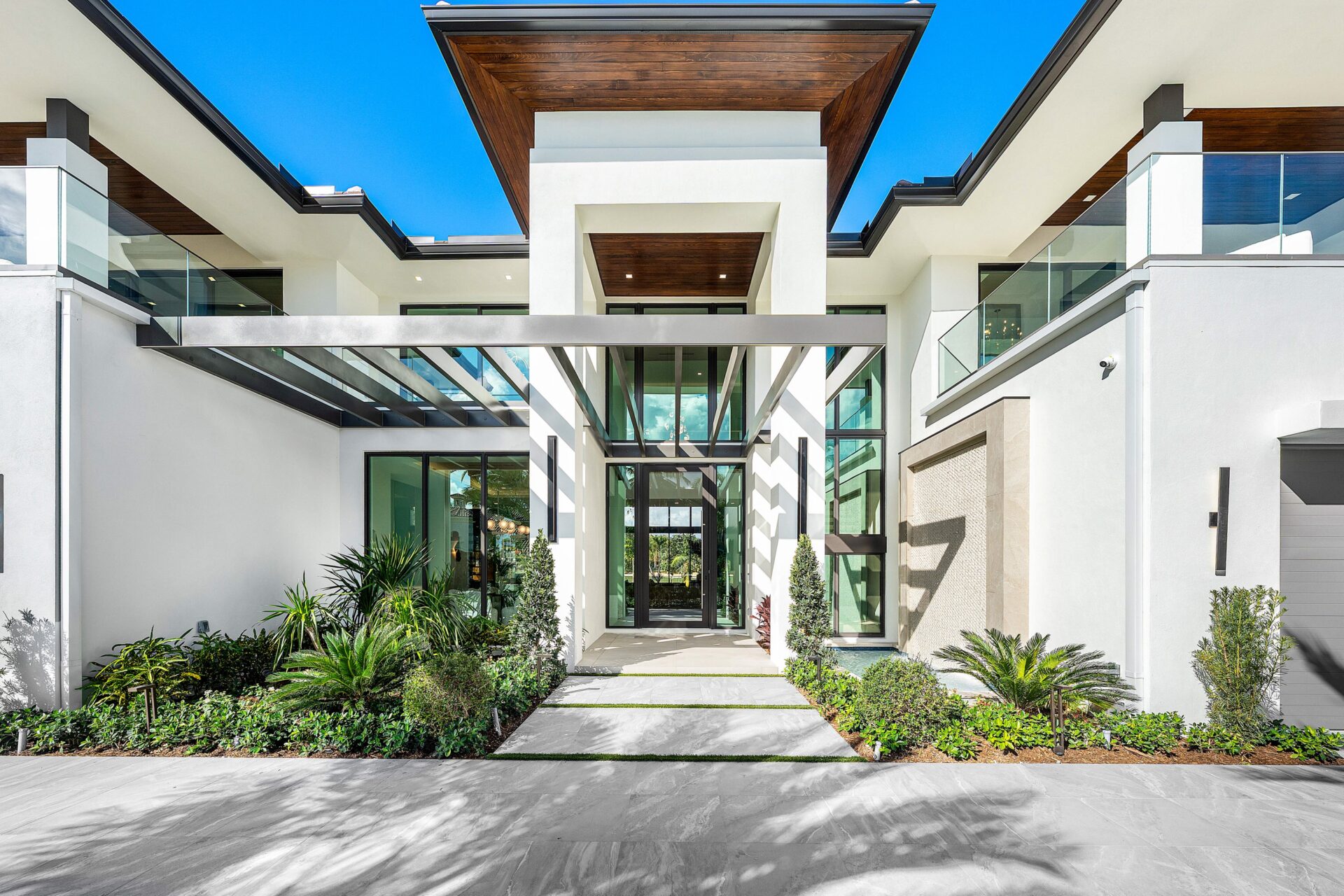 Large stone pavers lead through low tropical plant beds to a glass front door set beneath a steel trellis, which casts a grid of shadows across the entry.