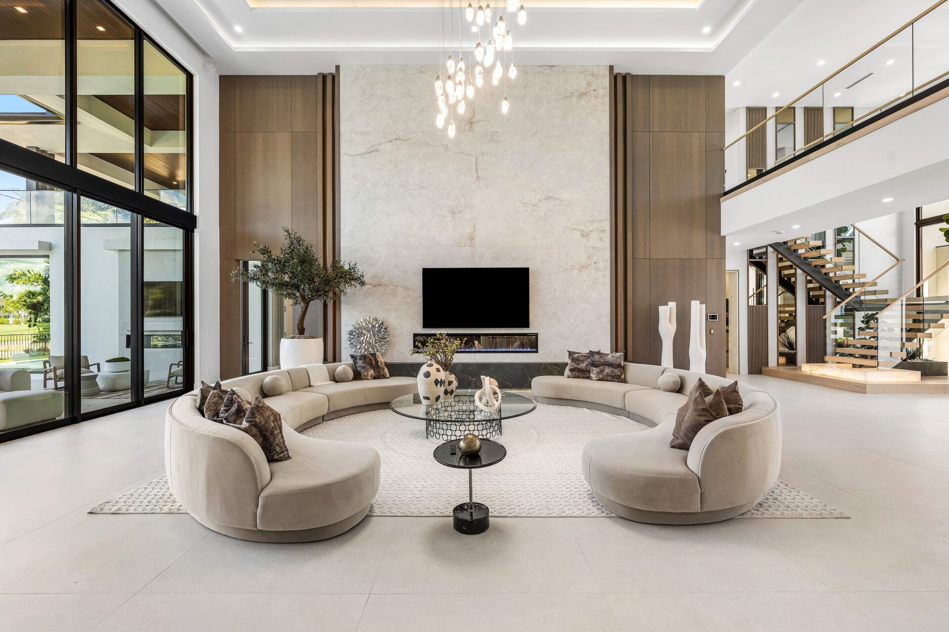 Curved sofas center the seating area beneath a hanging light fixture, while a tall stone fireplace and media wall anchor one side of the room.