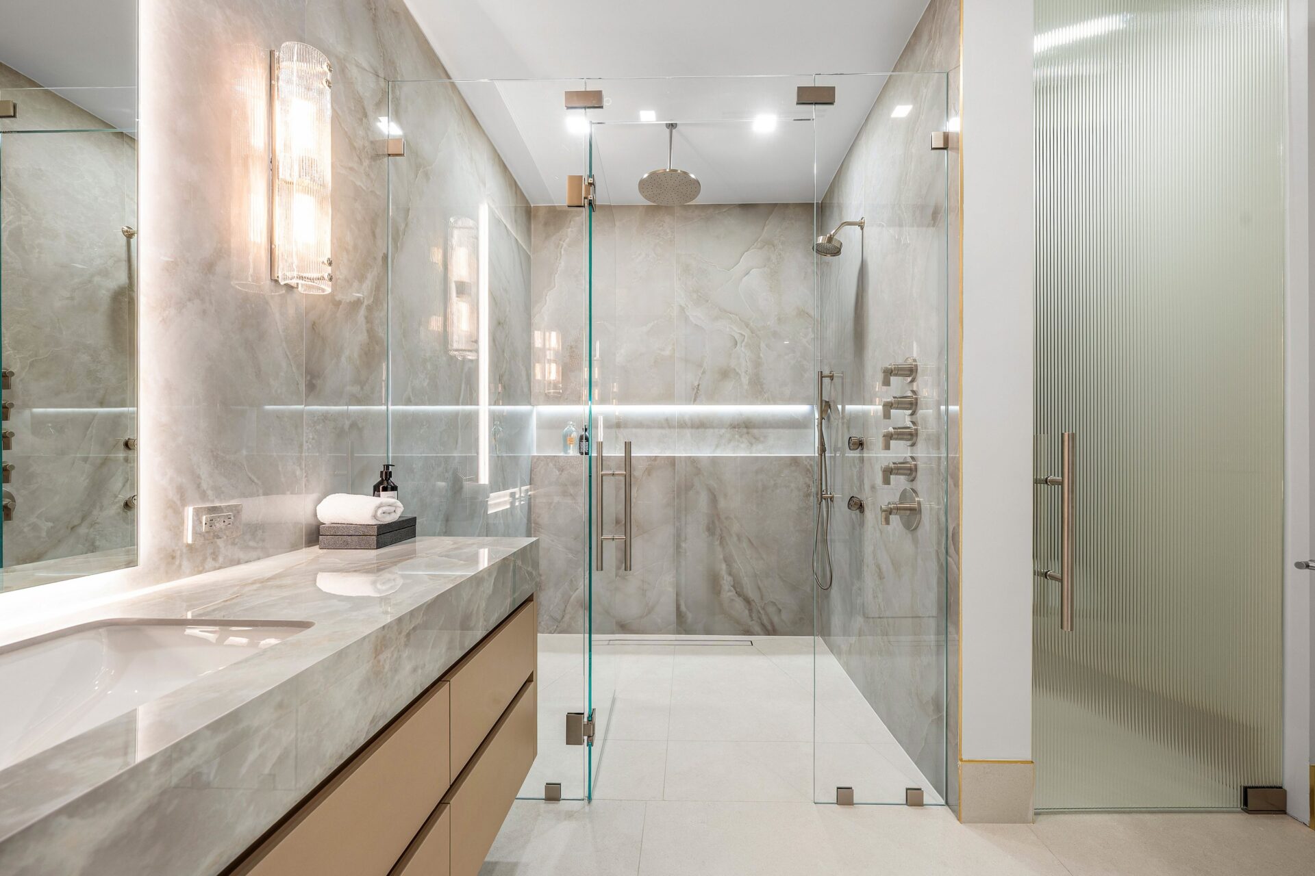 In the second bathroom, the shower becomes the main focal point, enclosed in glass and wrapped in full-height stone, with multiple controls and fixtures built into one wall.