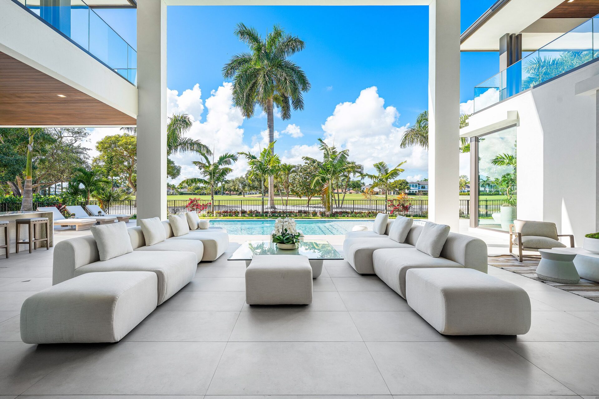 Large sectional seating sits beneath the double-height overhang, framed by the pool straight ahead and glass-lined balconies extending from the second level of the house.