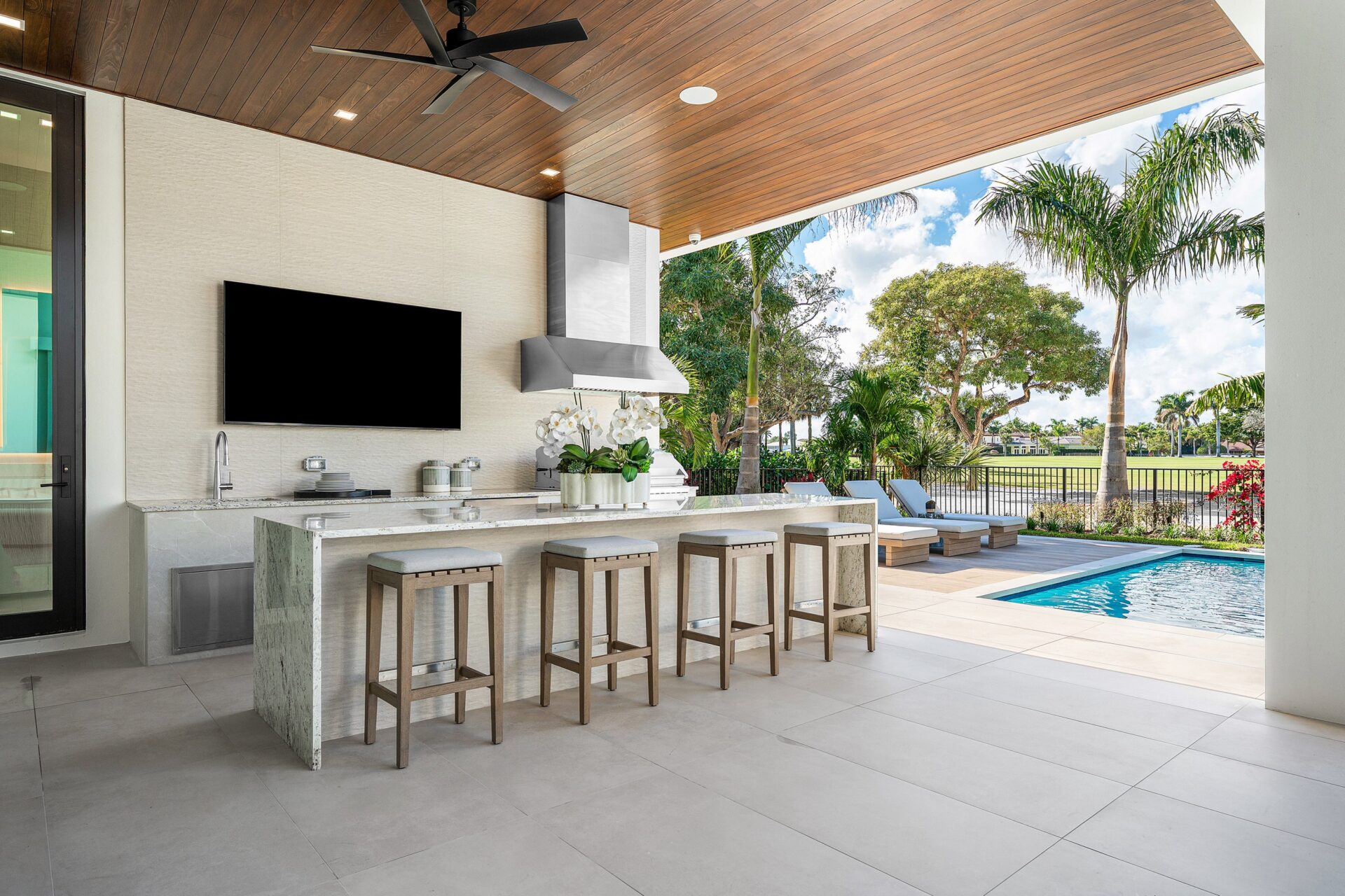 Off to one side, the outdoor kitchen is built into the covered terrace with a stone-topped bar, grill, sink, and wall-mounted television.