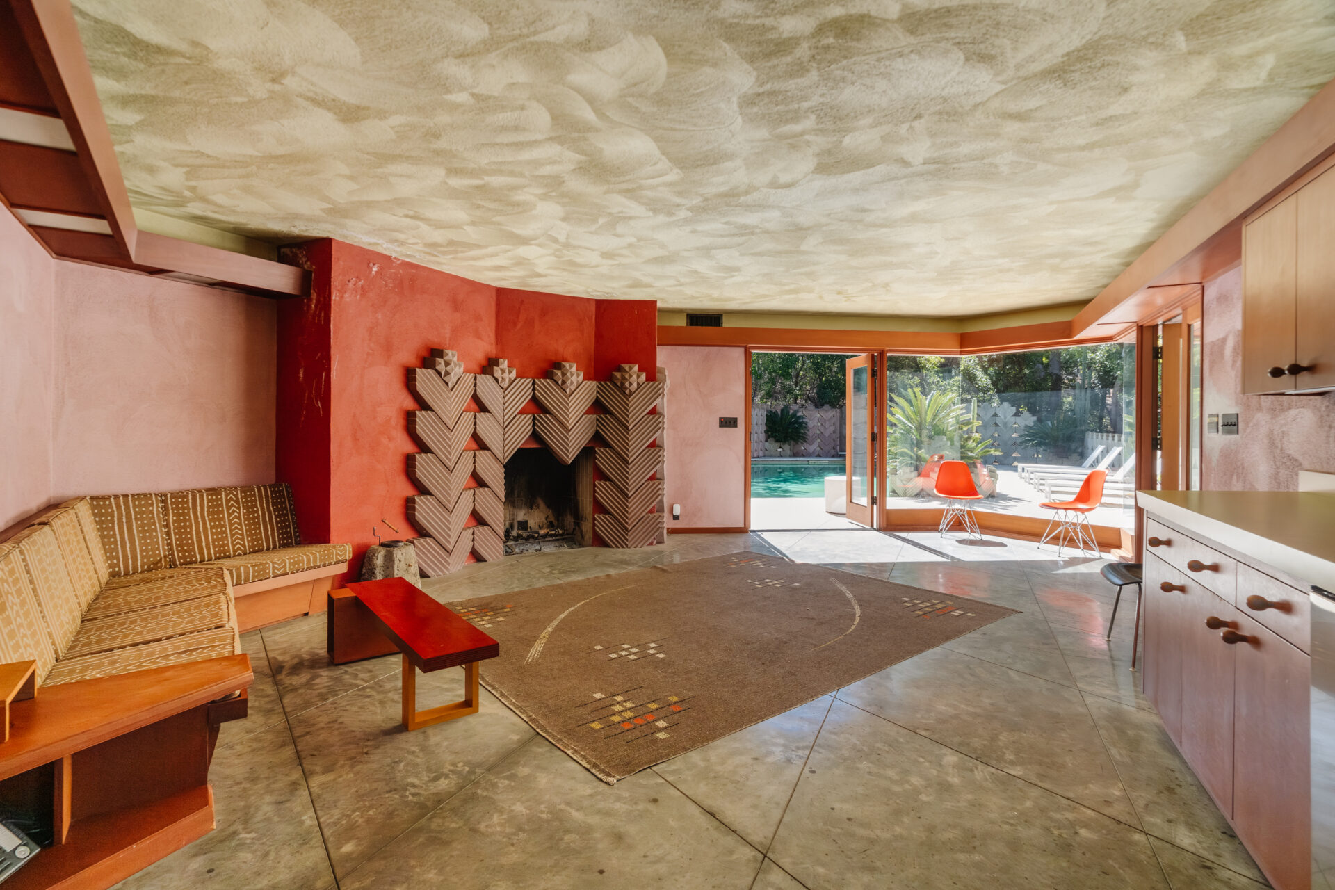 A sculptural fireplace framed by geometric tilework is set against warm red plaster walls and a textured ceiling that adds depth overhead. 