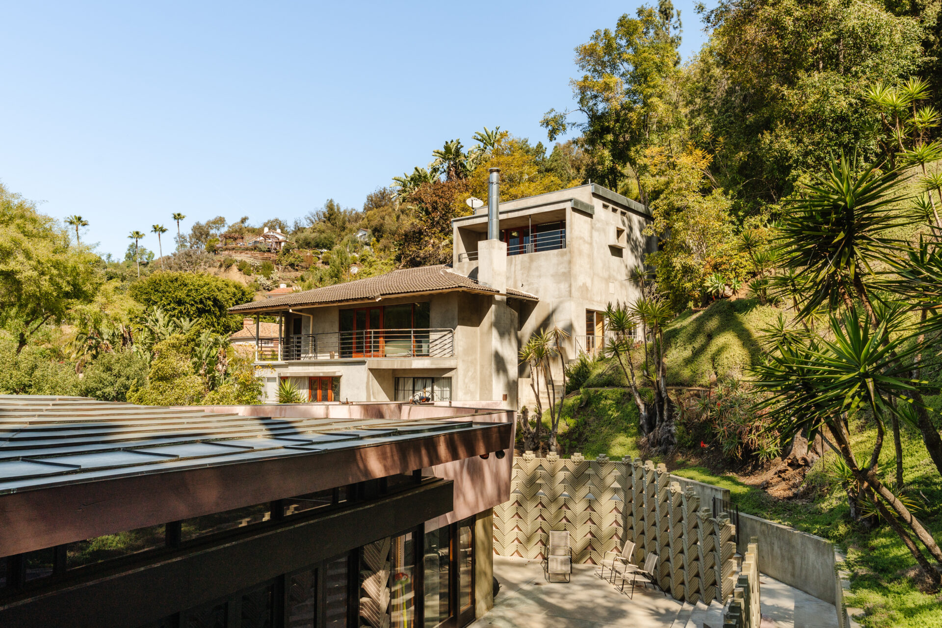 Large grey building rises above a one story home with lush trees surrounding it.