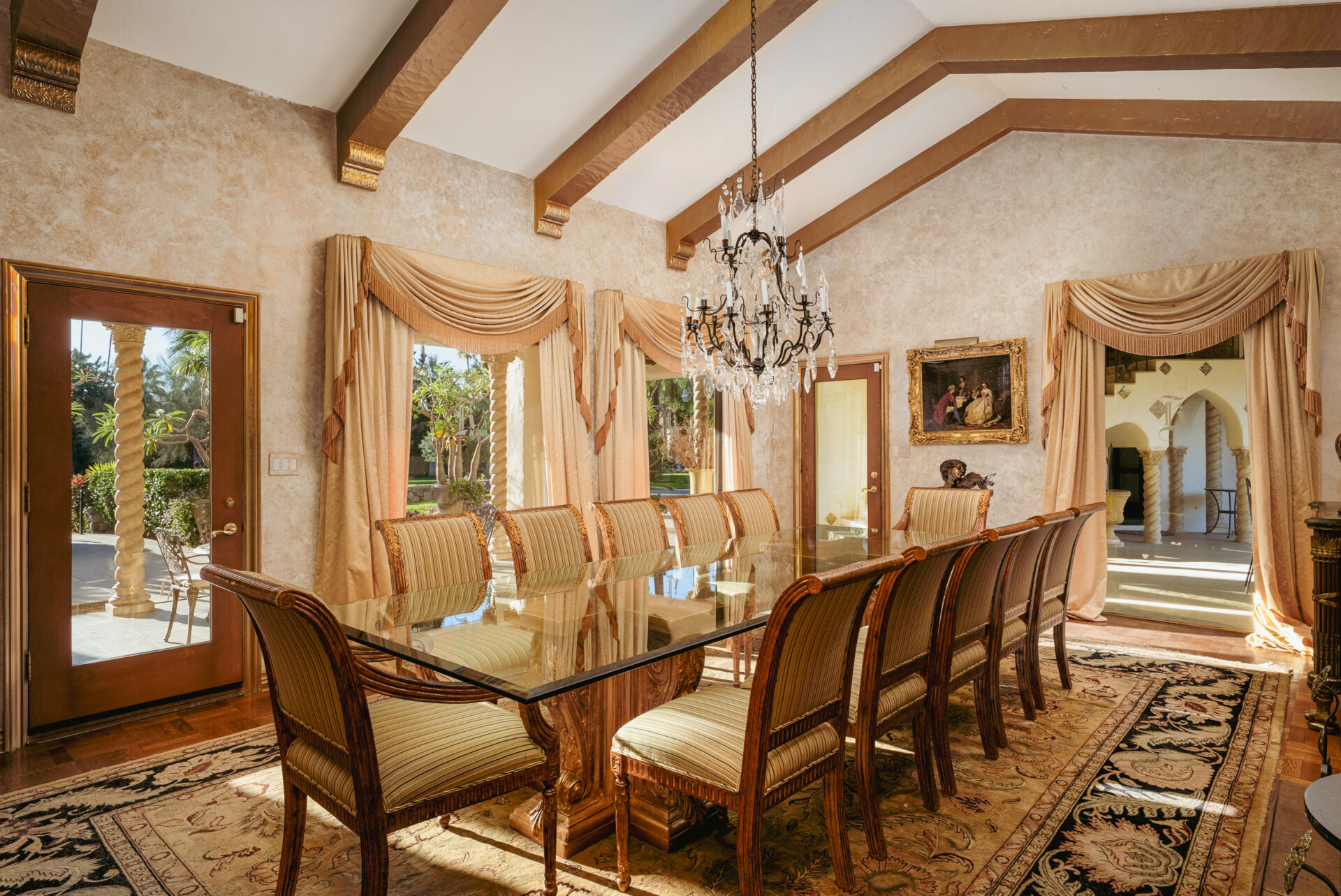 The formal dining room includes a long glass dining table surrounded by upholstered chairs that sit beneath a crystal chandelier.