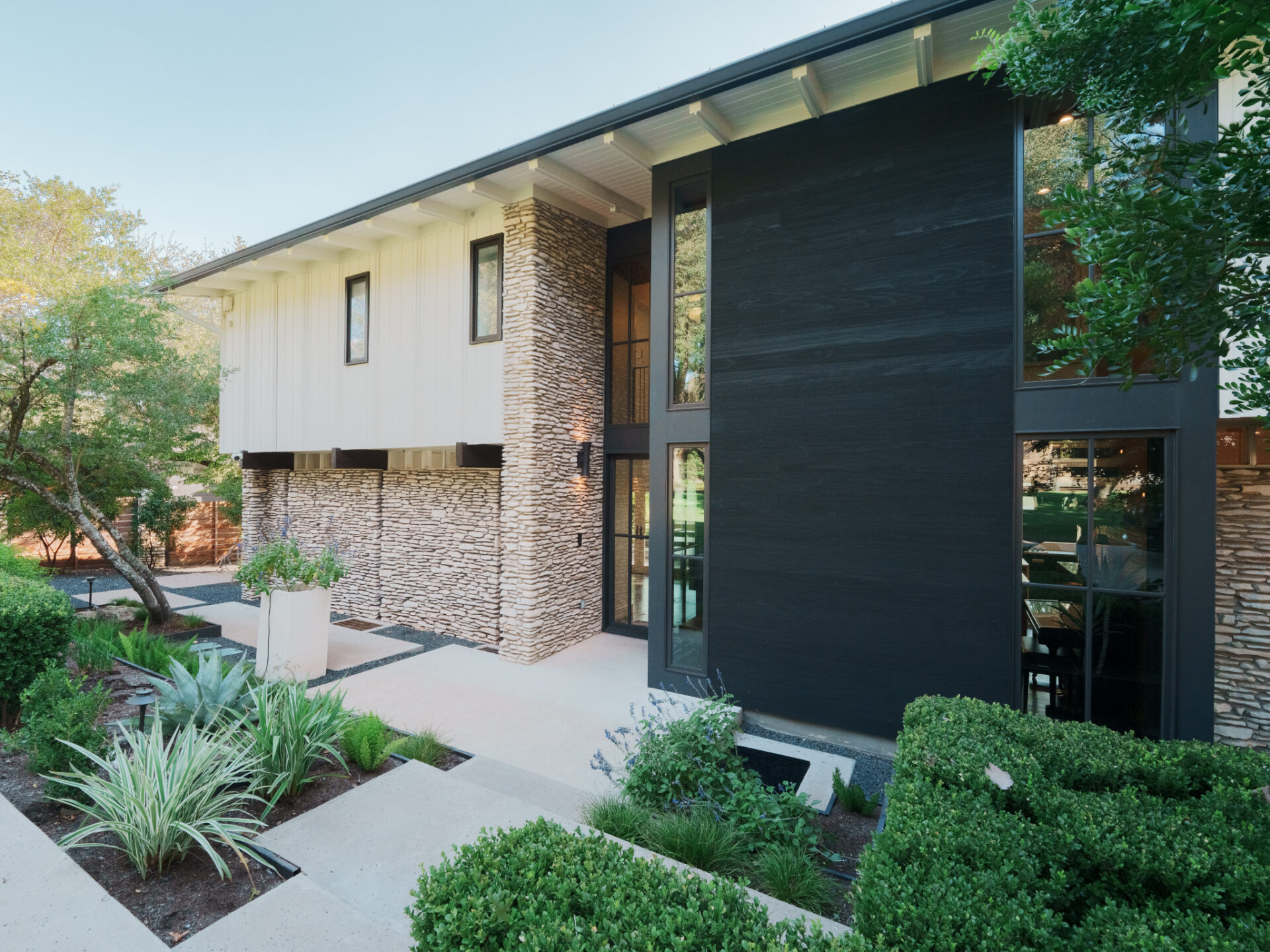 As you move closer, a mix of light-toned stone and crisp white siding wraps the exterior, contrasted by a bold vertical section of dark cladding that rises two stories at the center of the home. 