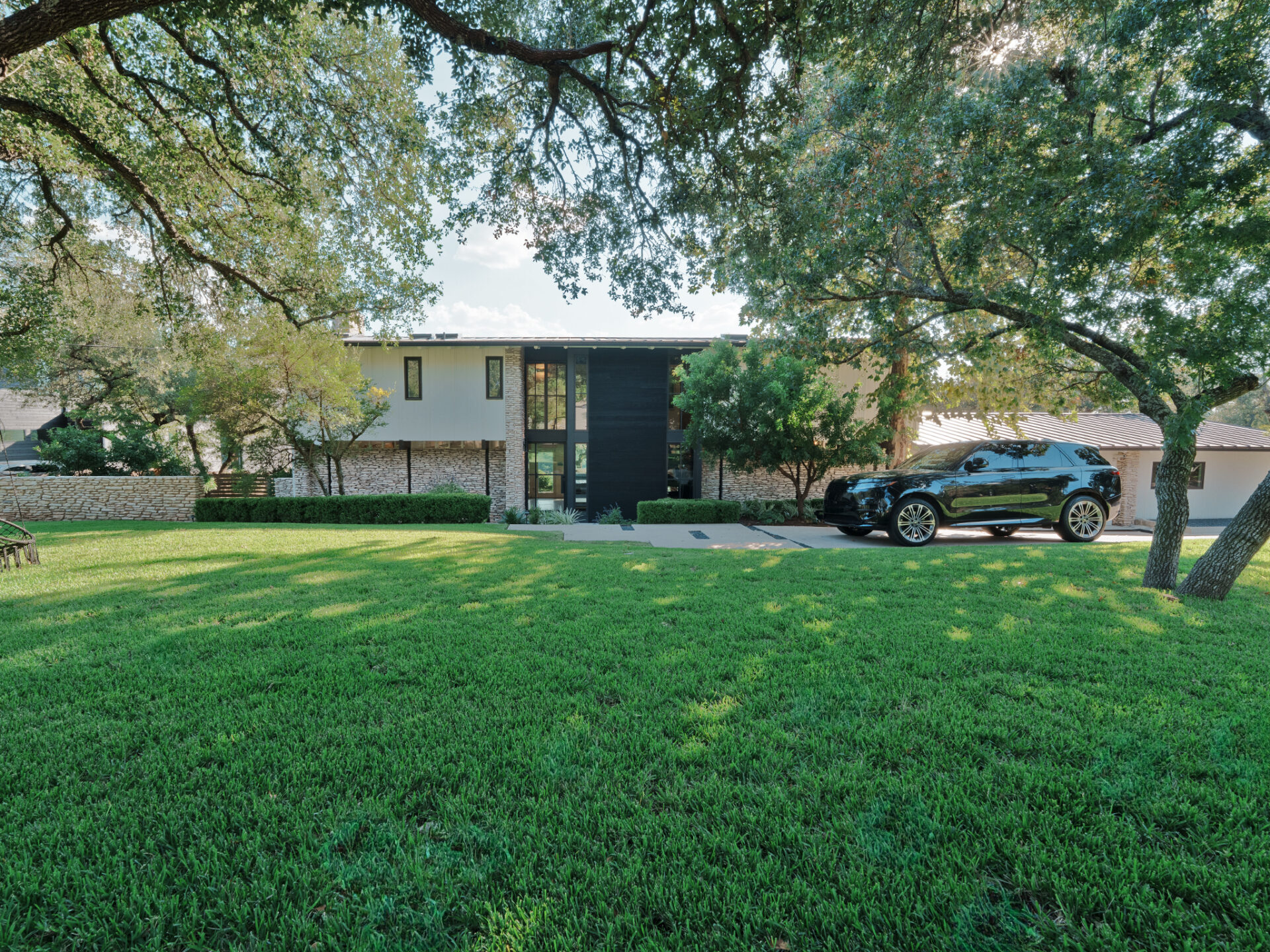 As you walk up to the house, a wide stretch of lawn unfolds beneath mature oak trees, their branches framing the exterior and casting patterned shadows across the grass and driveway. 