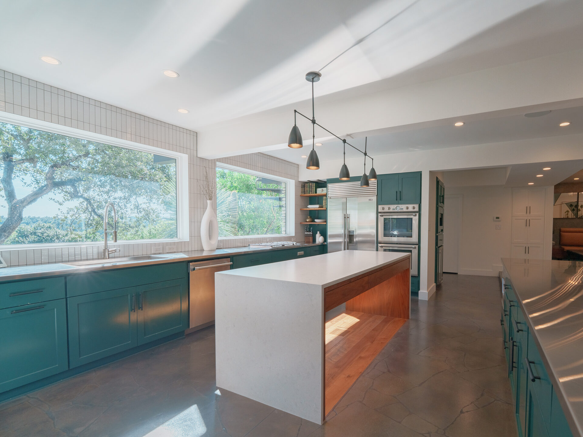 A stainless steel built-in refrigerator and double wall ovens are integrated into teal cabinetry that surrounds a large waterfall-edge island in the center of the room.
