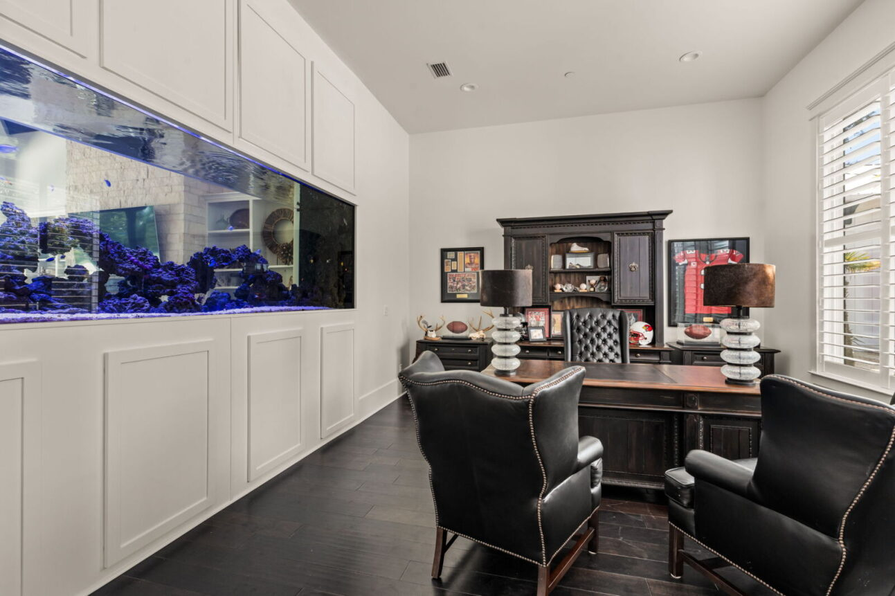 Inside the room, a traditional wood desk and leather chairs sit opposite the tank, with built-in cabinetry and framed memorabilia adding a more masculine, study-like atmosphere.