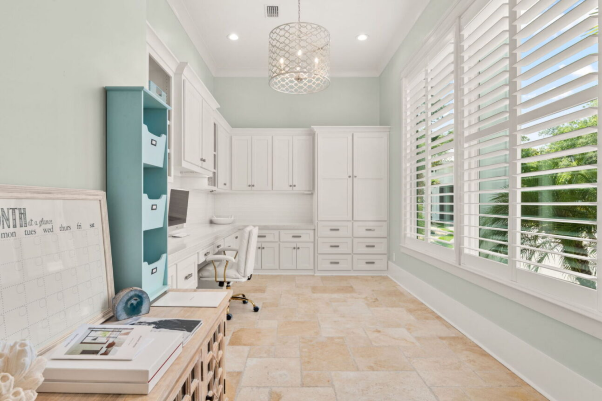 Soft green walls, tall plantation-shuttered windows, and white built-in cabinetry create a bright environment centered around a built-in desk and storage system.