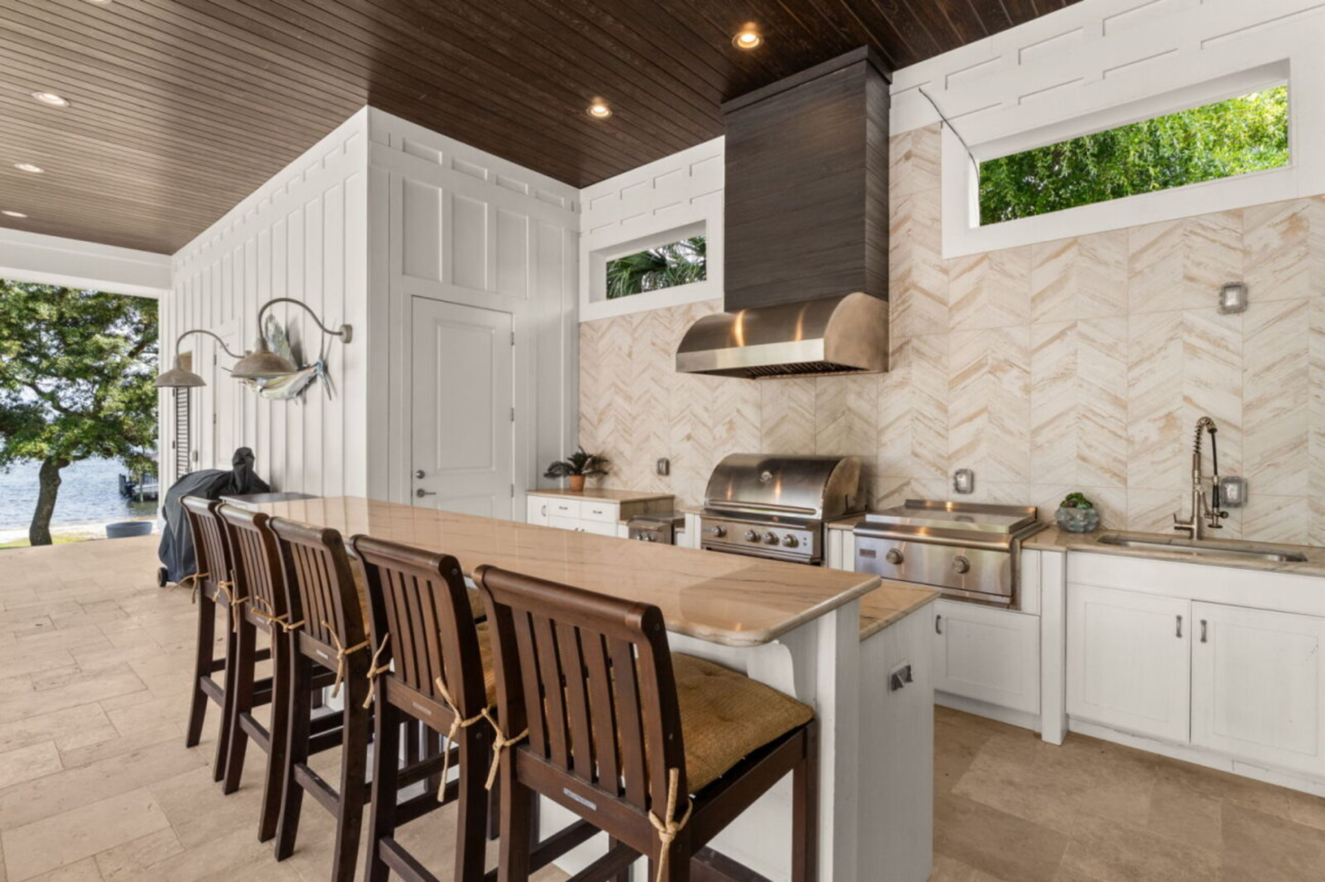 The space includes a full outdoor kitchen with a built-in grill, flat-top cooktop, sink, and prep space, along with a long bar counter lined with seating. 