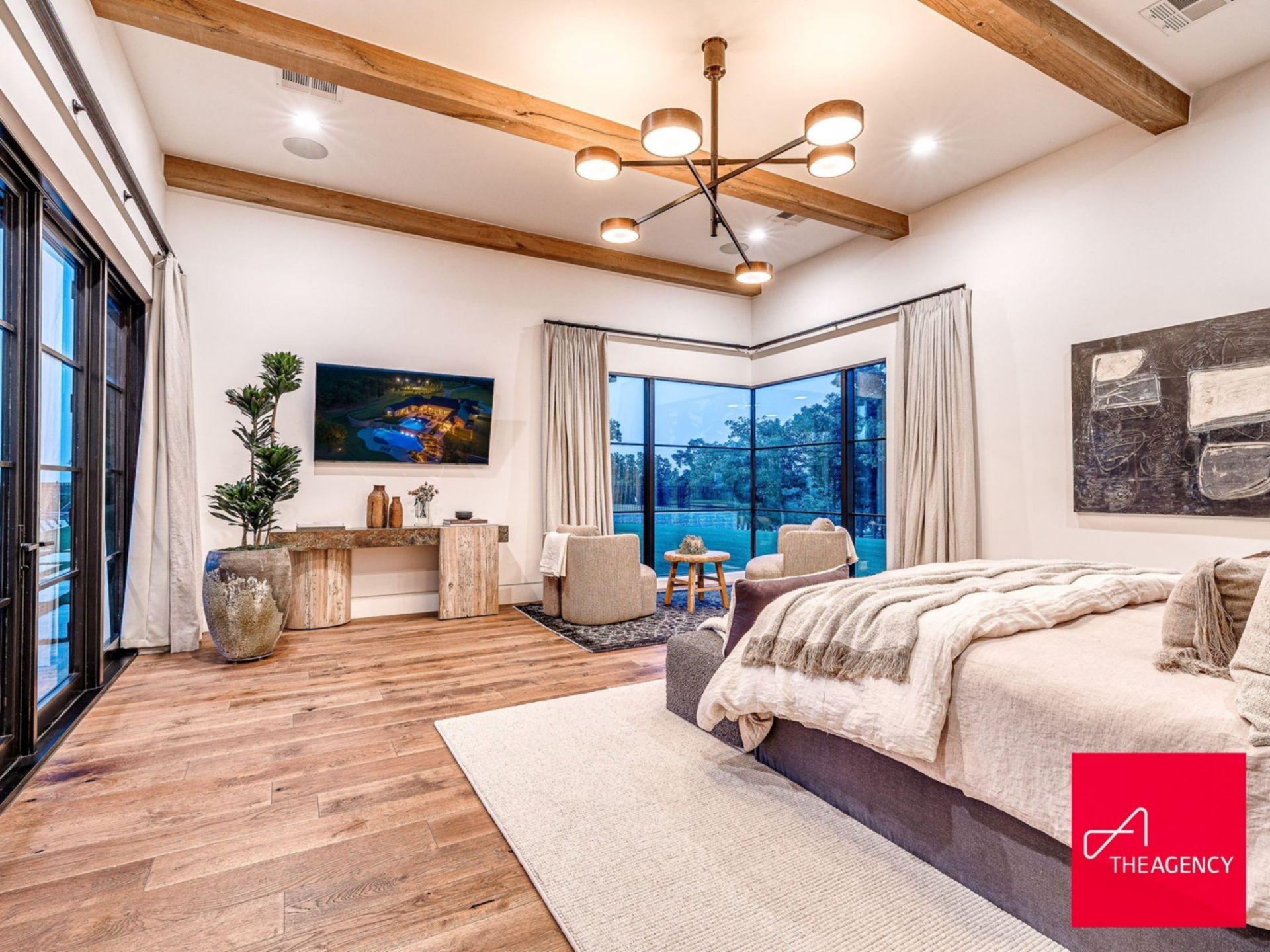 The bedroom itself sits beneath exposed wood beams that stretch across the ceiling, adding warmth overhead, while expansive corner windows bring in views of the surrounding acreage.