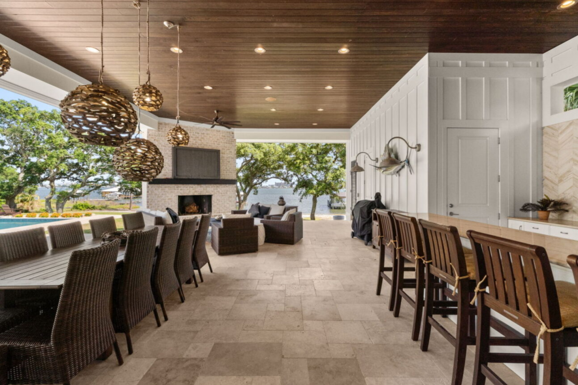 Next to the kitchen, a large dining table sits beneath woven pendant lights, while a lounge area gathers around a fireplace and television, all positioned to look out toward the water and pool.