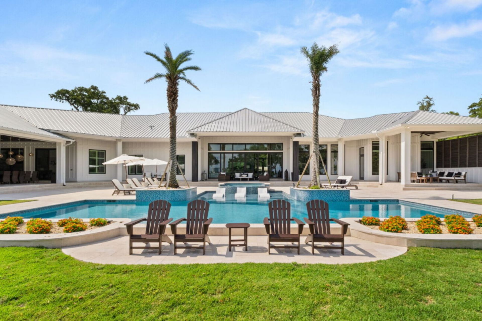 The pool is framed by wide stone decking and raised planters with palm trees, creating a symmetrical focal point in the backyard. 