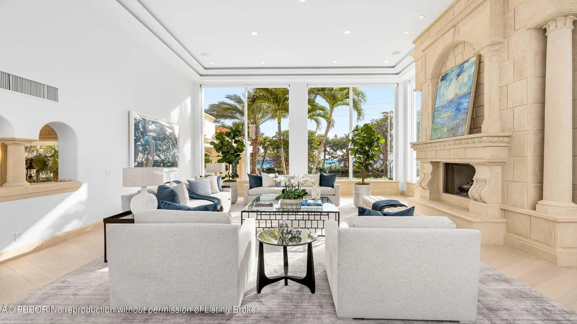 At the center, a symmetrical seating arrangement of soft white sofas and armchairs surrounds a glass-top coffee table, while a carved stone fireplace lines the room along one wall. 