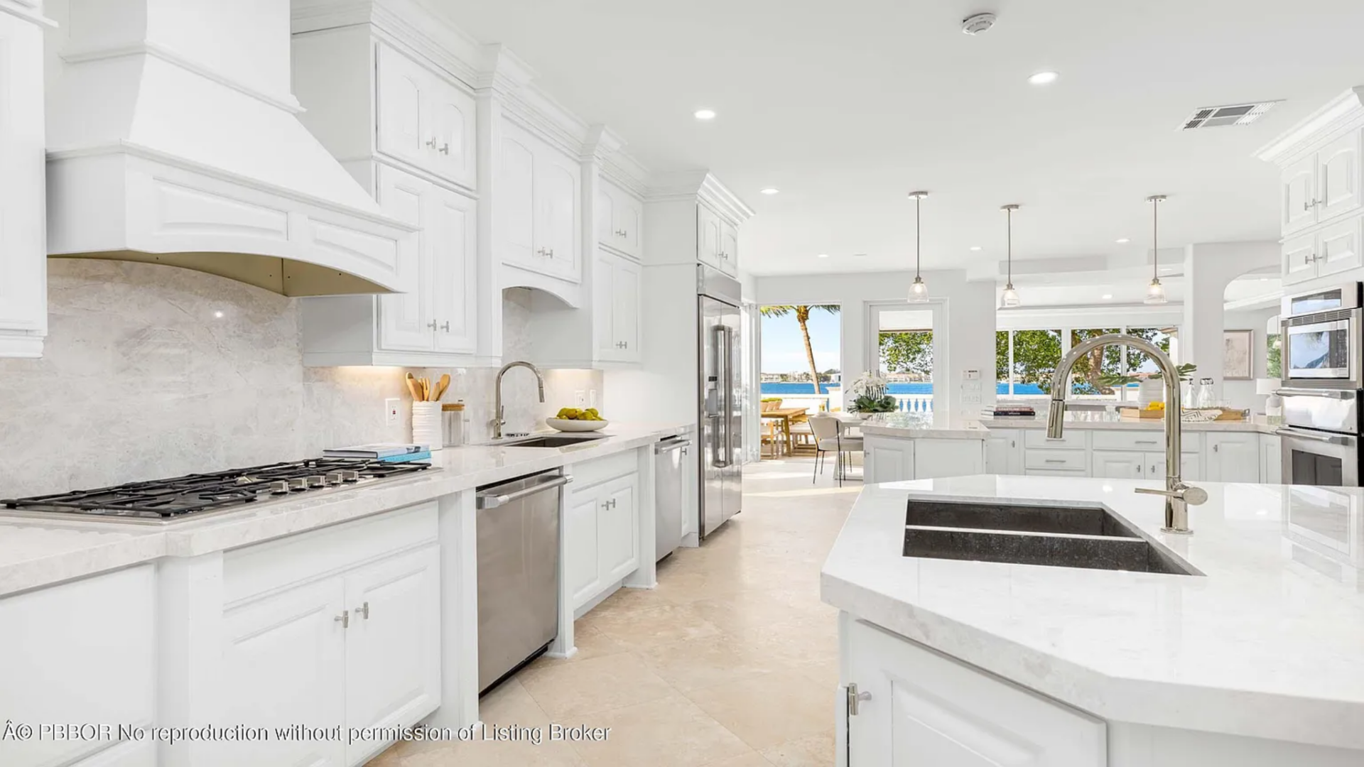 At the center sits a large island with a built-in sink and prep space, while stainless steel appliances, including a gas cooktop with a custom hood and double wall ovens, line the perimeter.